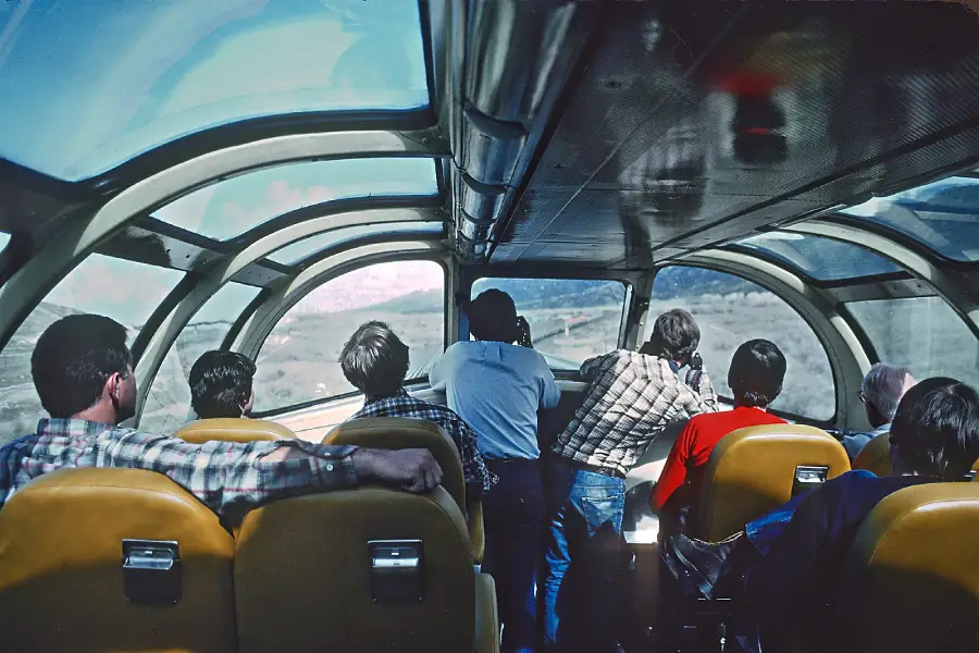 A group of people sit and look out the curved glass windows of a vintage train observation car, enjoying scenic views of the landscape passing by.