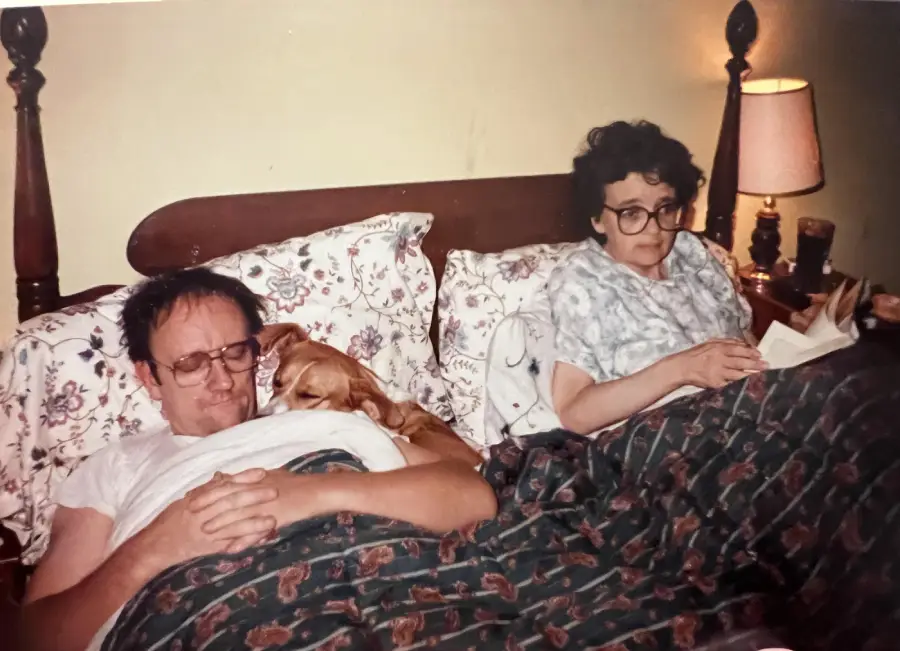 A man and a woman relax in bed with a dog between them. The man appears to be resting while the woman reads a book. Both have glasses, and a lamp is on the nightstand beside them.