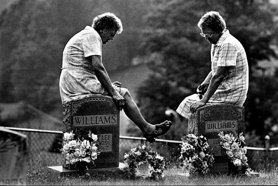 Two elderly women sit facing each other on gravestones labeled “Williams,” surrounded by flowers, in a grassy outdoor cemetery.