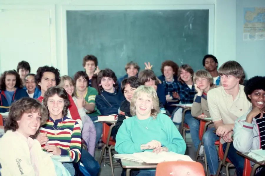 A group of smiling students sit at desks in a classroom, posing for a photo. A chalkboard is in the background, and some students make playful gestures. Everyone appears happy and engaged.