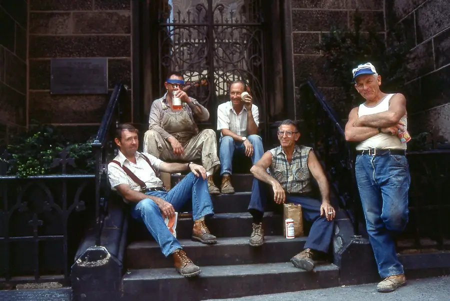 Five men in work clothes relax on stone steps, some holding drinks. They appear to be construction or utility workers, with casual postures and expressions, in front of a wrought-iron gate and brick building.
