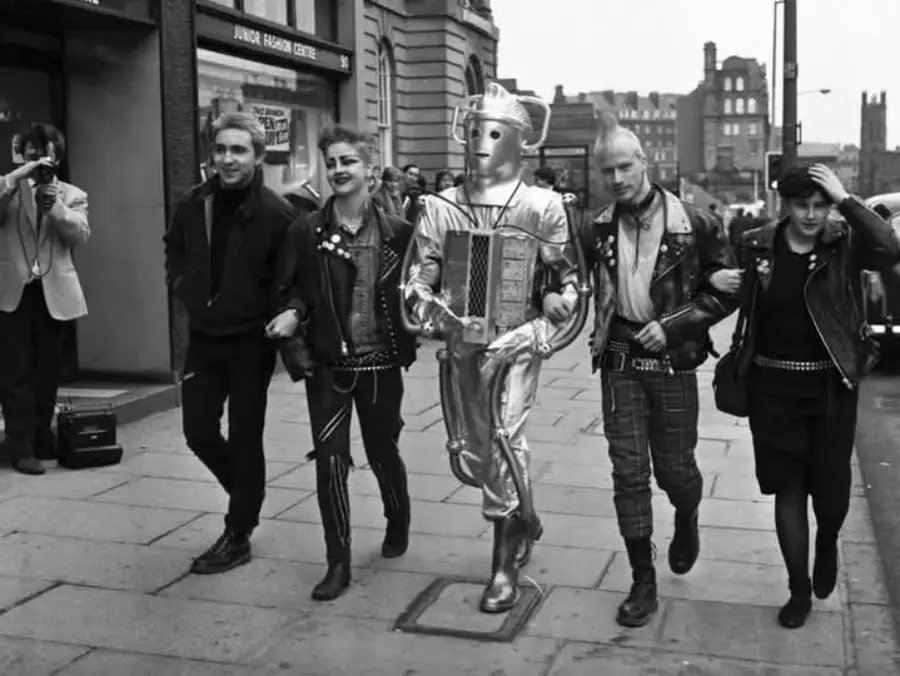 Four people in punk clothing and a person in a robot costume walk arm-in-arm down a city sidewalk, while a photographer captures the moment.
