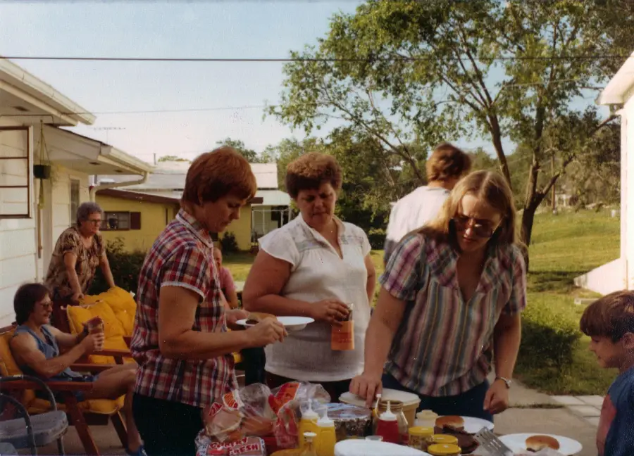 People gather outdoors near a table with food, preparing plates at what appears to be a casual backyard gathering. Houses and trees are visible in the background, with several people seated and others serving themselves.