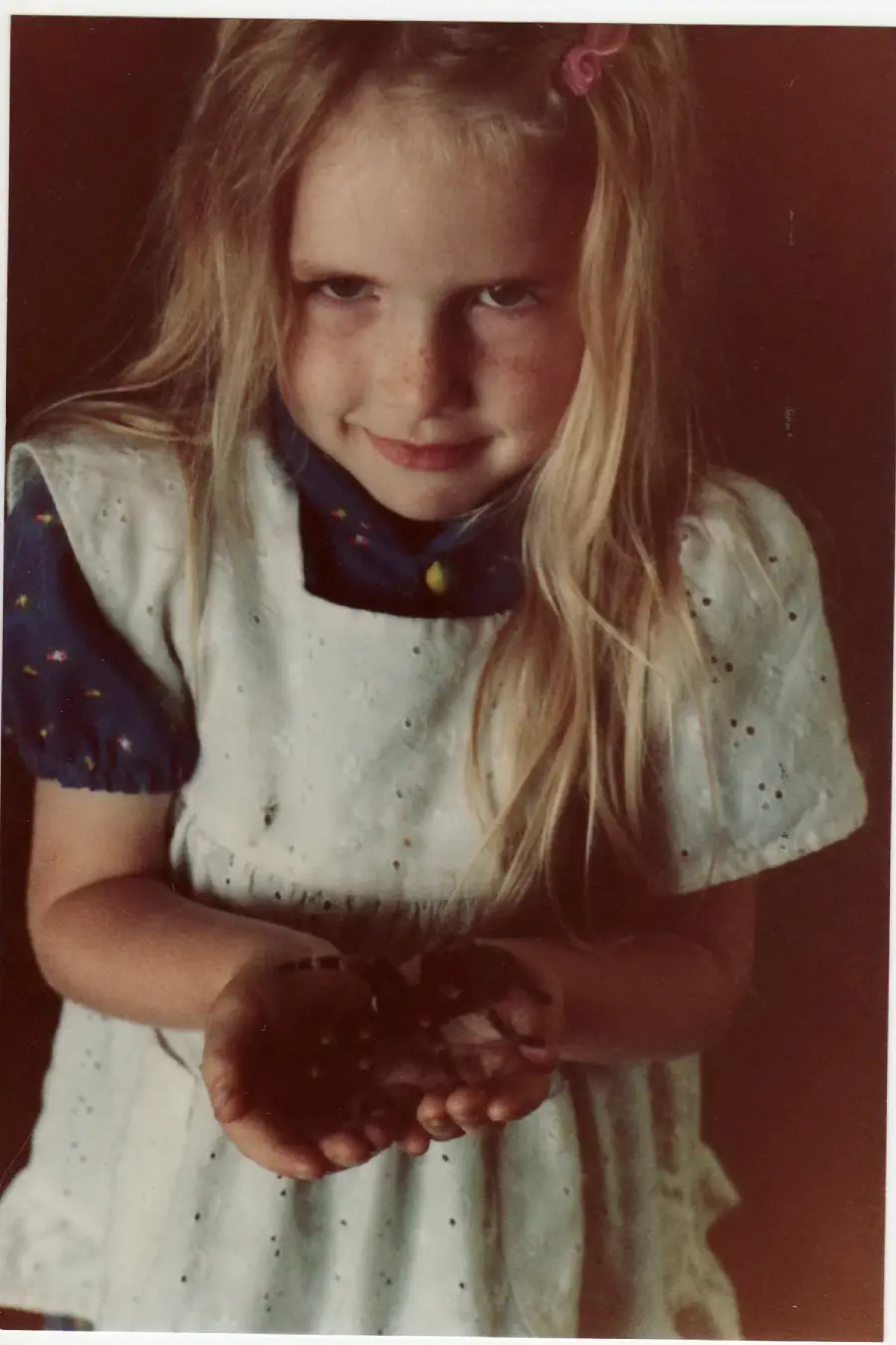 A young girl with long blonde hair and a white dress holds something small in her cupped hands, smiling slightly and looking up at the camera with a playful expression.