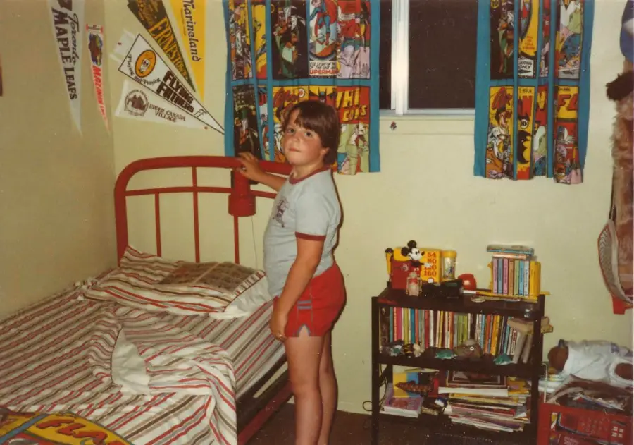 A young boy in a vintage bedroom stands beside a red bed, touching a wall lamp. The room features superhero curtains, sports pennants, books, and toys on a shelf, with striped bedding on the bed.