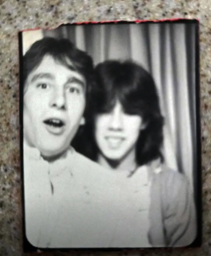 Black and white photo booth picture of two young people smiling, with one person in front making a surprised face. A curtain is in the background and the photo rests on a speckled surface.