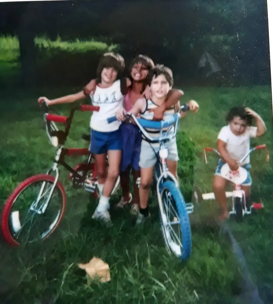 Four children pose outdoors; three stand together with bikes, smiling, while one younger child sits on a tricycle nearby, touching their head. The background is grassy and green with trees.