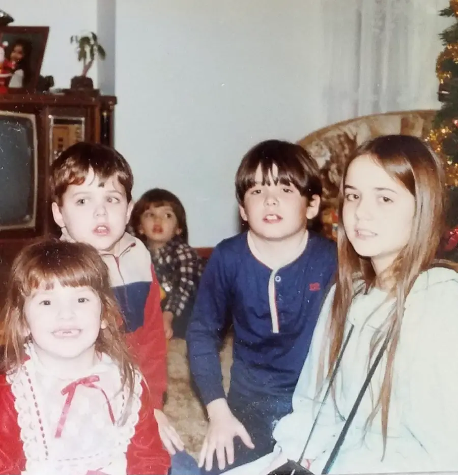 Four children sit together near a Christmas tree in a living room, with one more child in the background. There’s an old television and framed photo on a cabinet. The kids appear to be posing for a holiday photo.