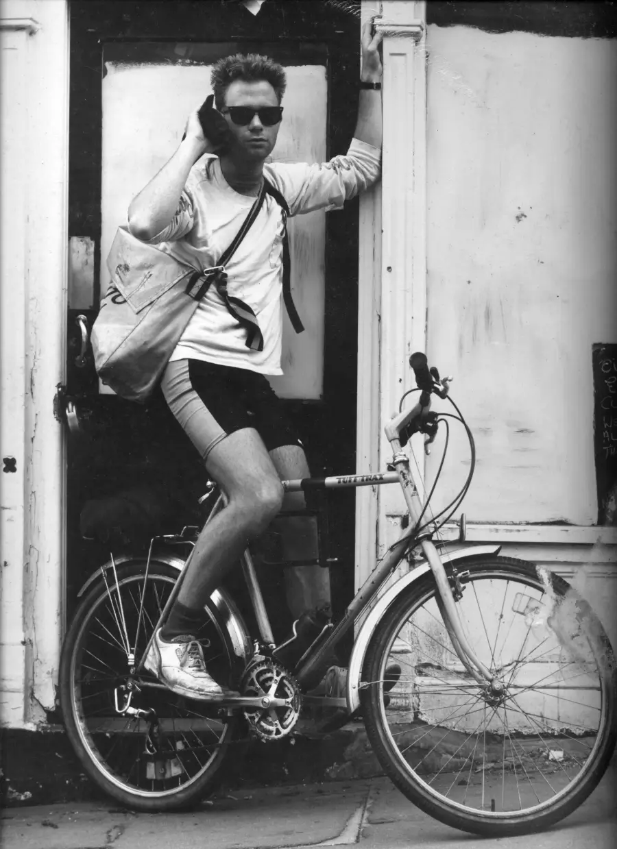 A man wearing sunglasses leans against a doorway while sitting on a bicycle, holding a phone to his ear and carrying a shoulder bag. The scene is in black and white, and the man appears relaxed and casual.