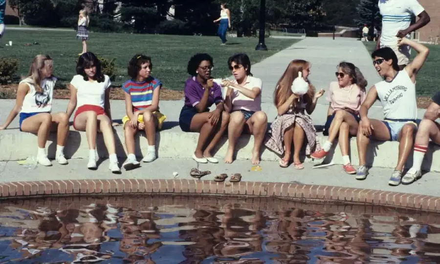 A group of eight people sit by a small fountain, chatting and eating snacks on a sunny day. They appear relaxed and are dressed in casual summer clothes. Some have their feet in the water.