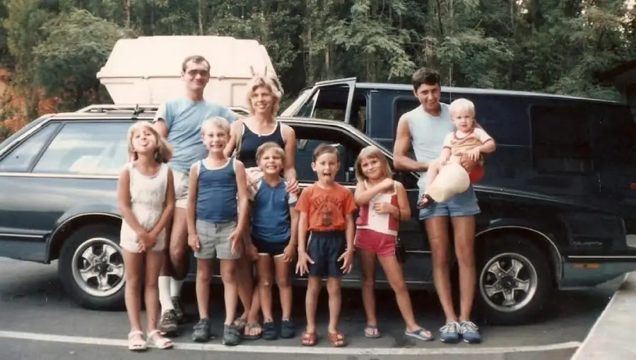 Eight children and two adults pose and smile in front of a dark station wagon, with trees in the background. One adult is holding a toddler, and several kids wear shorts and tank tops.