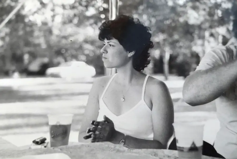 A woman with short curly hair, wearing a light tank top, sits at a picnic table outdoors. She looks to the side, with drinks and a blurred figure next to her. The photo is black and white.
