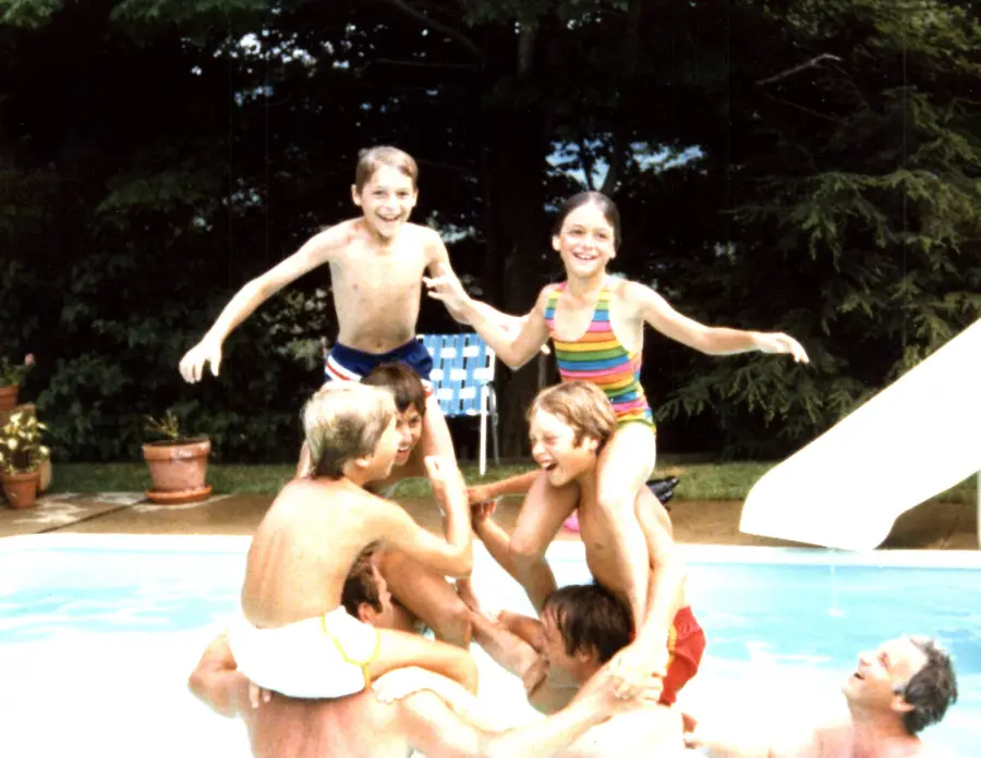 Kids in colorful swimsuits are playing in a pool, forming a human pyramid with two children on top, all smiling and having fun. There are trees and a slide in the background.