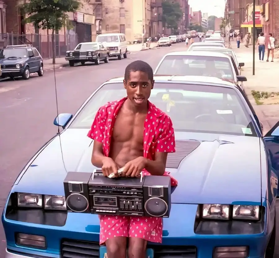 A young man in a red patterned outfit and open shirt sits on the hood of a blue car, holding a boombox on his lap, on a city street lined with parked cars.