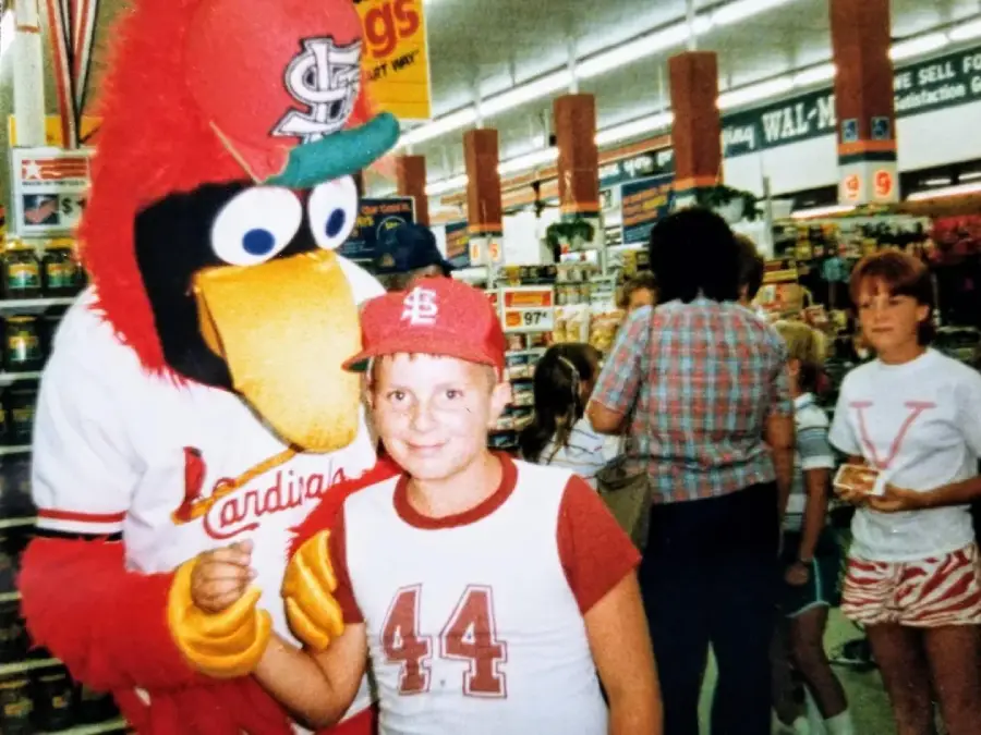 A child in a Cardinals shirt and cap smiles next to a person in a large red bird mascot costume inside a brightly lit store. Other shoppers and store shelves are visible in the background.