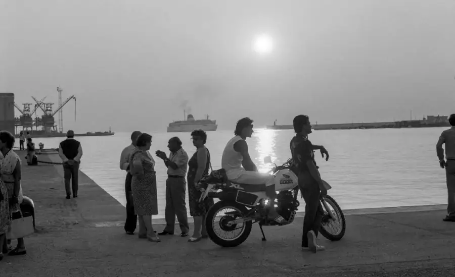 People stand and talk near a dock at sunset. A motorcycle is parked nearby, and a ship is visible on the water in the background. The scene is in black and white.
