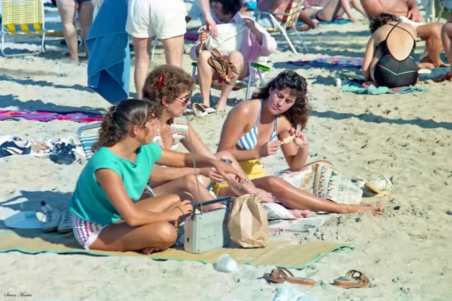 Three women sit on towels at a crowded beach, talking and eating. One holds a radio, another wears a striped bikini, and people relax in the background. Sand, sun, and summer vibes fill the scene.