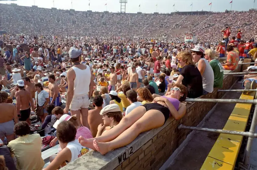A woman in sunglasses lies on a stadium railing among a large crowd of people, many wearing summer clothes, during what appears to be a concert or outdoor event on a sunny day.