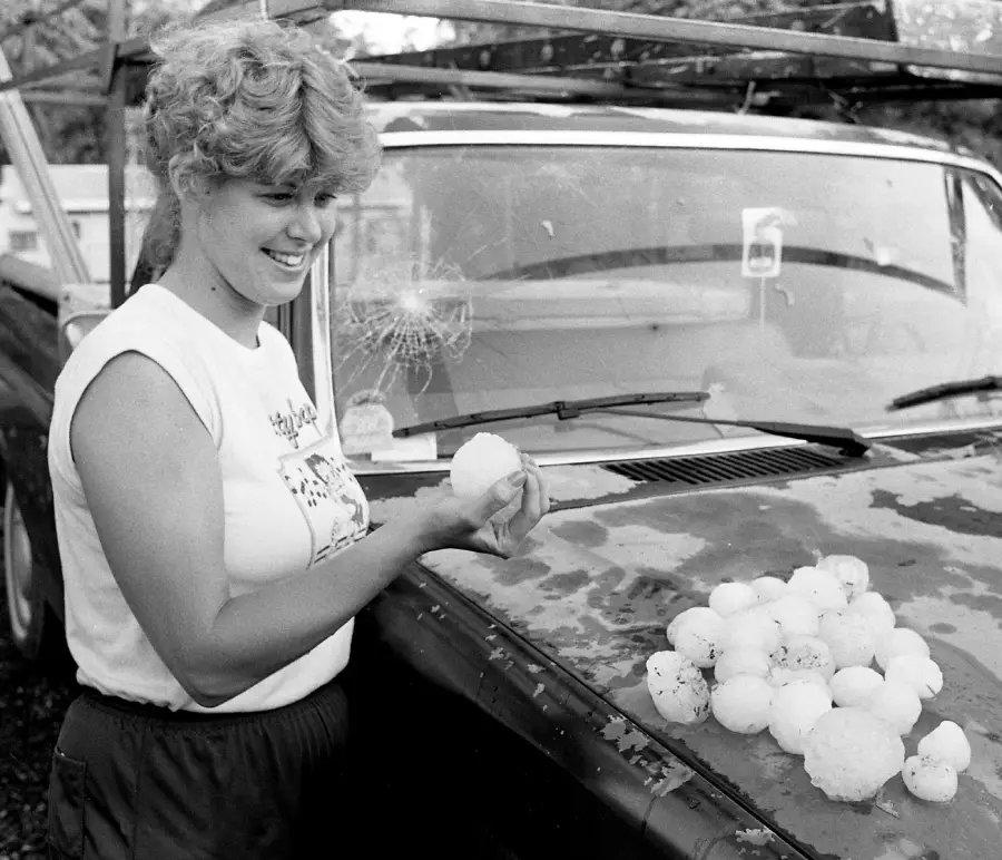 A woman smiles while holding large hailstones next to a car with a cracked windshield. More hailstones are piled on the car’s hood. The scene appears outdoors, and the image is black and white.