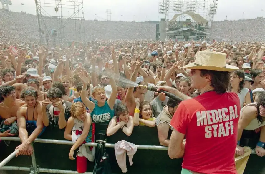 A person in a “Medical Staff” shirt sprays water on a large, excited crowd at an outdoor concert on a sunny day.