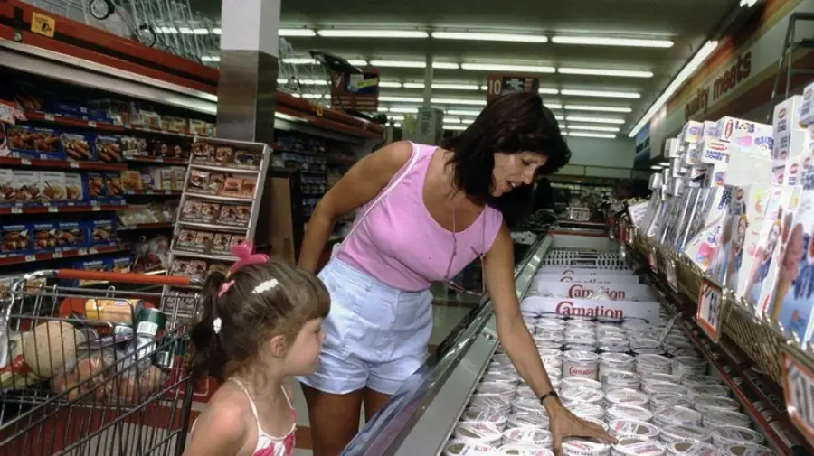 A woman in a pink tank top and white shorts selects items from a freezer in a grocery store while a young girl stands nearby with a shopping cart full of groceries.