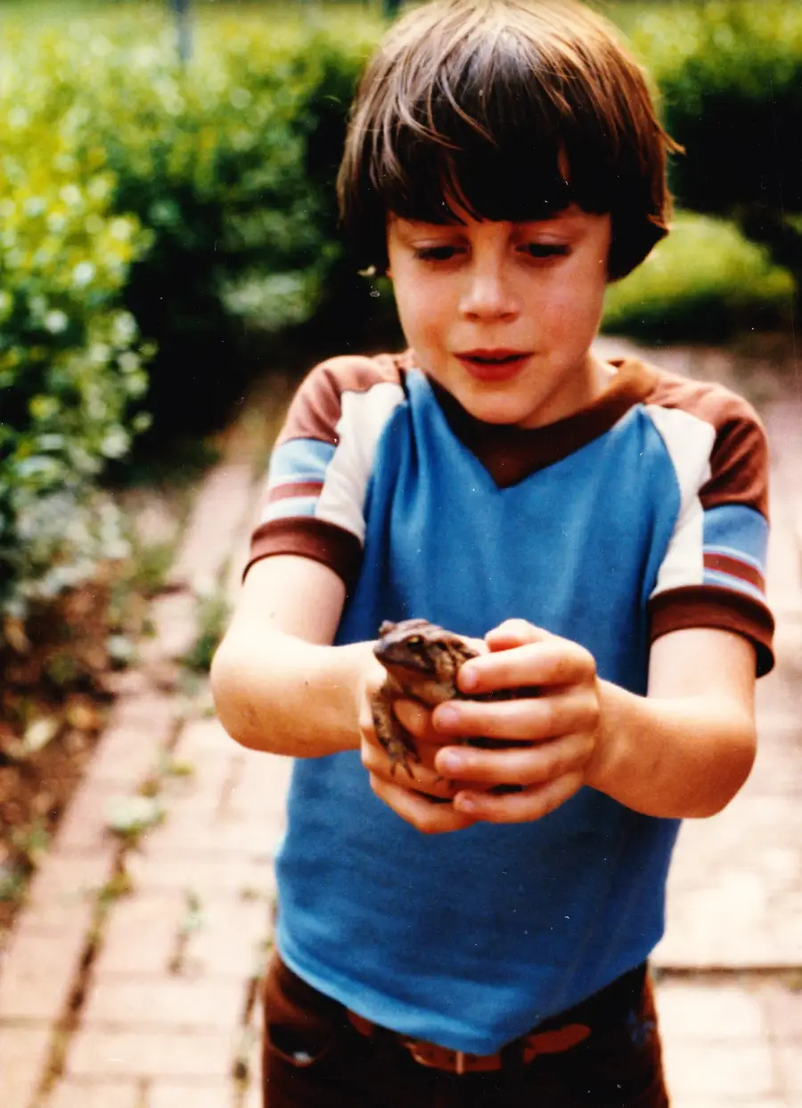 A young boy with brown hair, wearing a blue and brown shirt, stands on a brick path outside, smiling as he gently holds a small frog in both hands. Greenery is visible in the background.