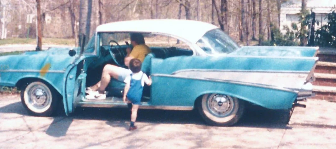 A child in blue overalls stands by the open door of a vintage teal car, while an adult sits inside. Trees and a house are visible in the background on a sunny day.