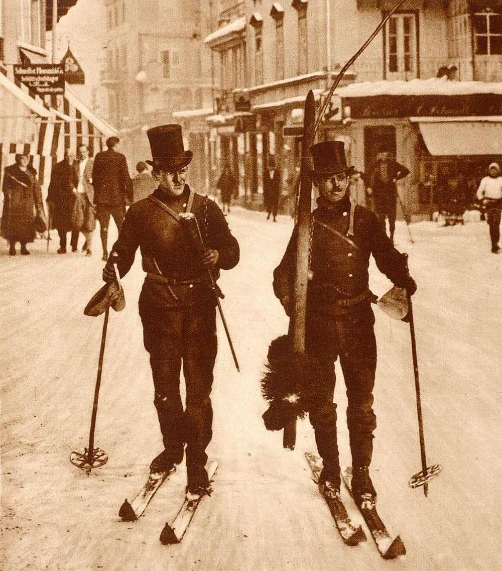 Two men in vintage clothing and top hats ski on a snowy street in an old town, carrying their poles and skis, while other people walk in the background among historic buildings.