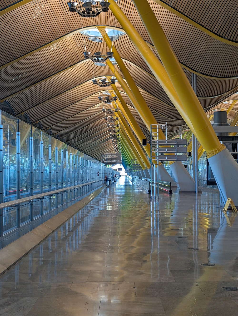 Spacious airport terminal with a high, curved wooden ceiling, yellow support columns, glass walls, and reflections on the shiny floor. Few people are visible in the distance.