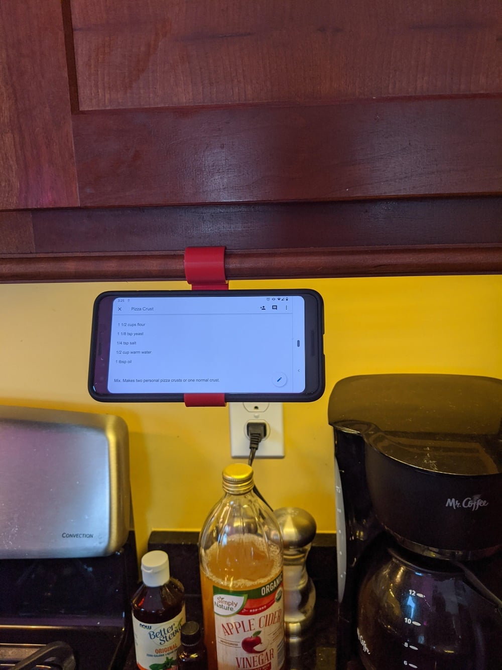 A phone displaying a recipe is clipped under a kitchen cabinet above a counter with coffee maker, apple cider vinegar, and other kitchen items.