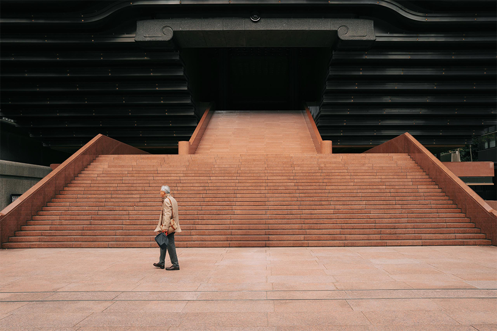 An older man in a beige jacket walks past wide, empty terracotta steps leading up to a large, modern building with dark horizontal architectural elements.