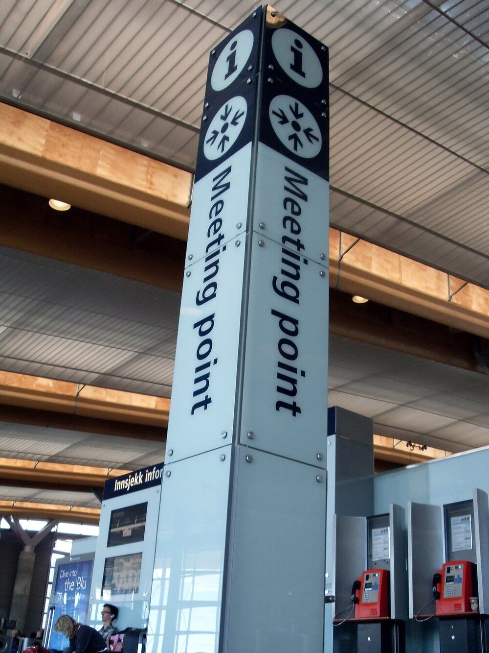 A tall sign in an airport reads “Meeting point” with information symbols. The background shows part of the terminal ceiling and red public telephones on the right.