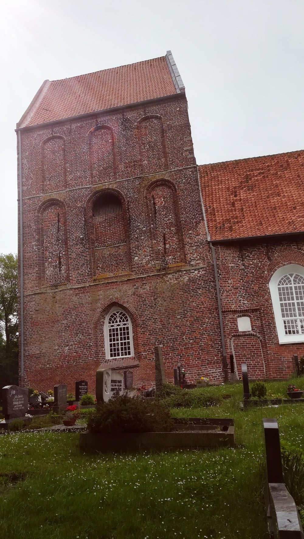 A historic brick church with a slanted tower, red-tiled roof, and arched windows stands behind a grassy graveyard with various tombstones on a cloudy day.