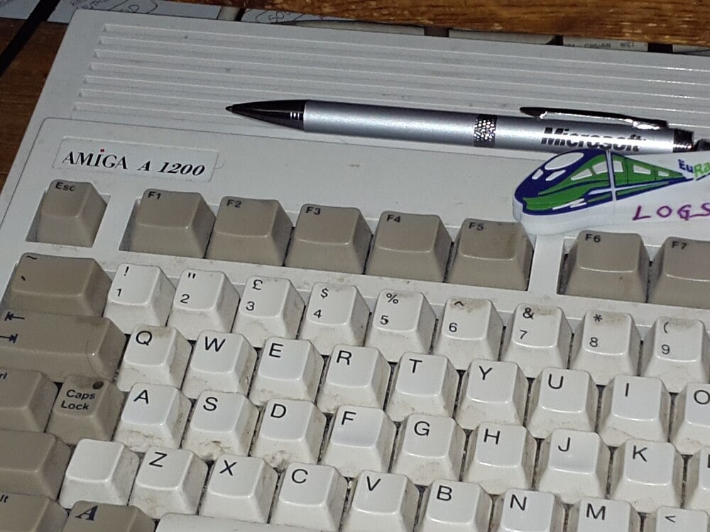 A close-up of an Amiga A1200 computer keyboard with a silver pen and a small sticker featuring a green train and the word “LOGS” placed on top.