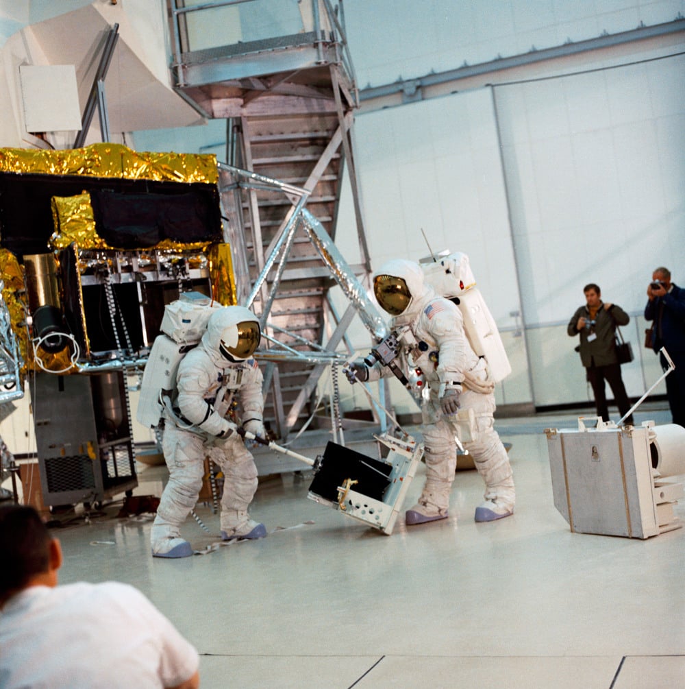 Two astronauts in spacesuits handle equipment in a brightly lit room with machinery and onlookers in the background, possibly during a training or demonstration.