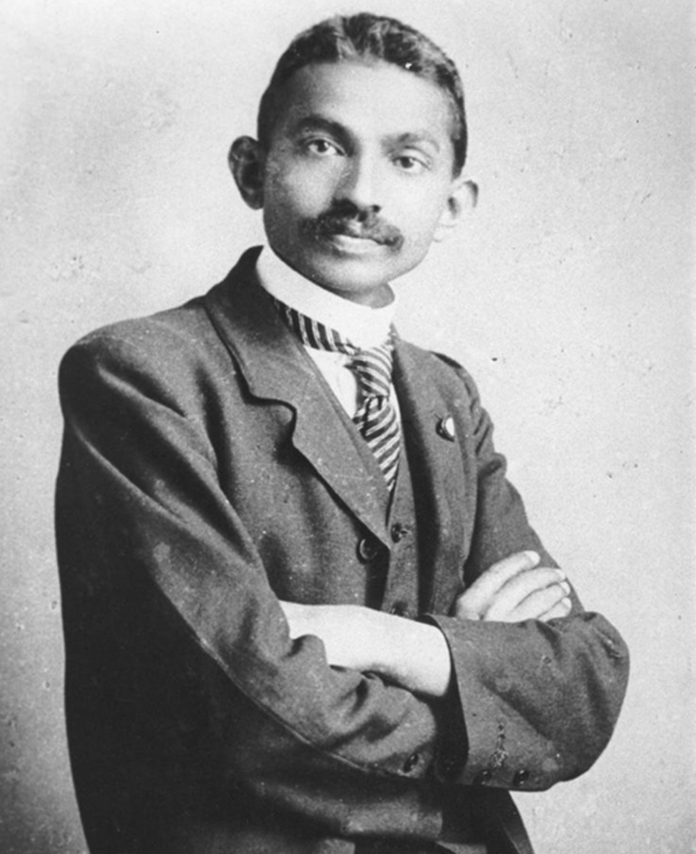 A young man with short hair and a mustache stands with arms crossed, wearing a suit, tie, and high-collared shirt, looking confidently at the camera in a black-and-white photograph.