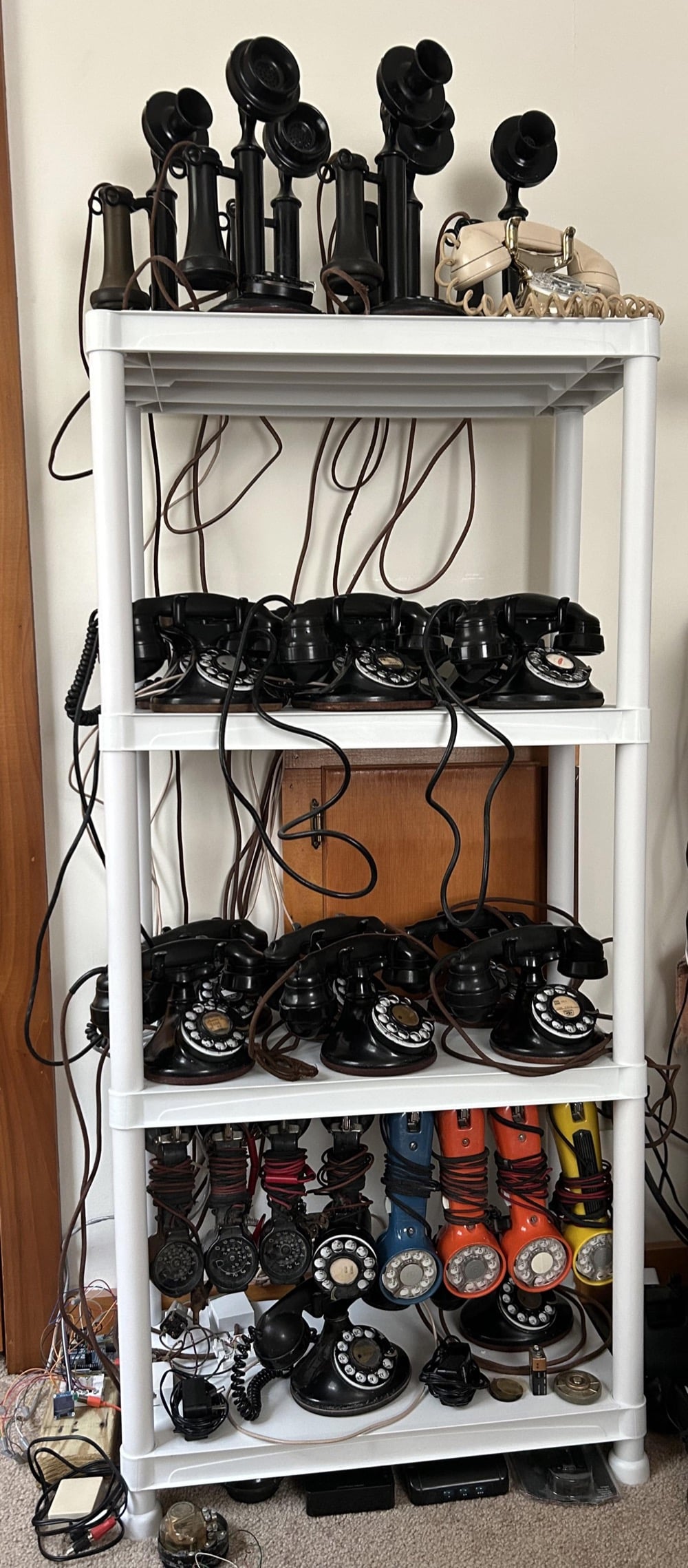 A white shelf holds a collection of vintage rotary phones and candlestick telephones, with cords hanging down. The phones are mostly black, with a few colorful models on the bottom shelf.