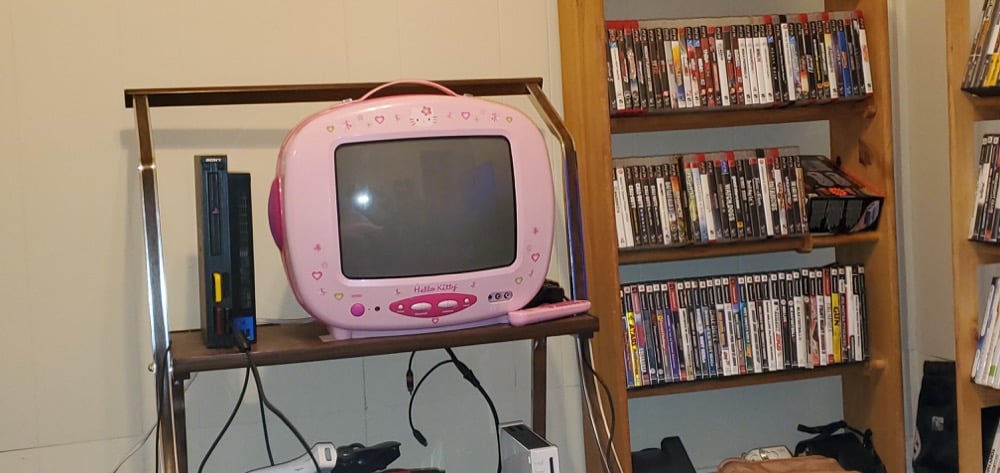 A pink Hello Kitty TV sits on a small stand beside a PlayStation 2 and gaming controllers. Behind them are shelves filled with video game and movie cases.