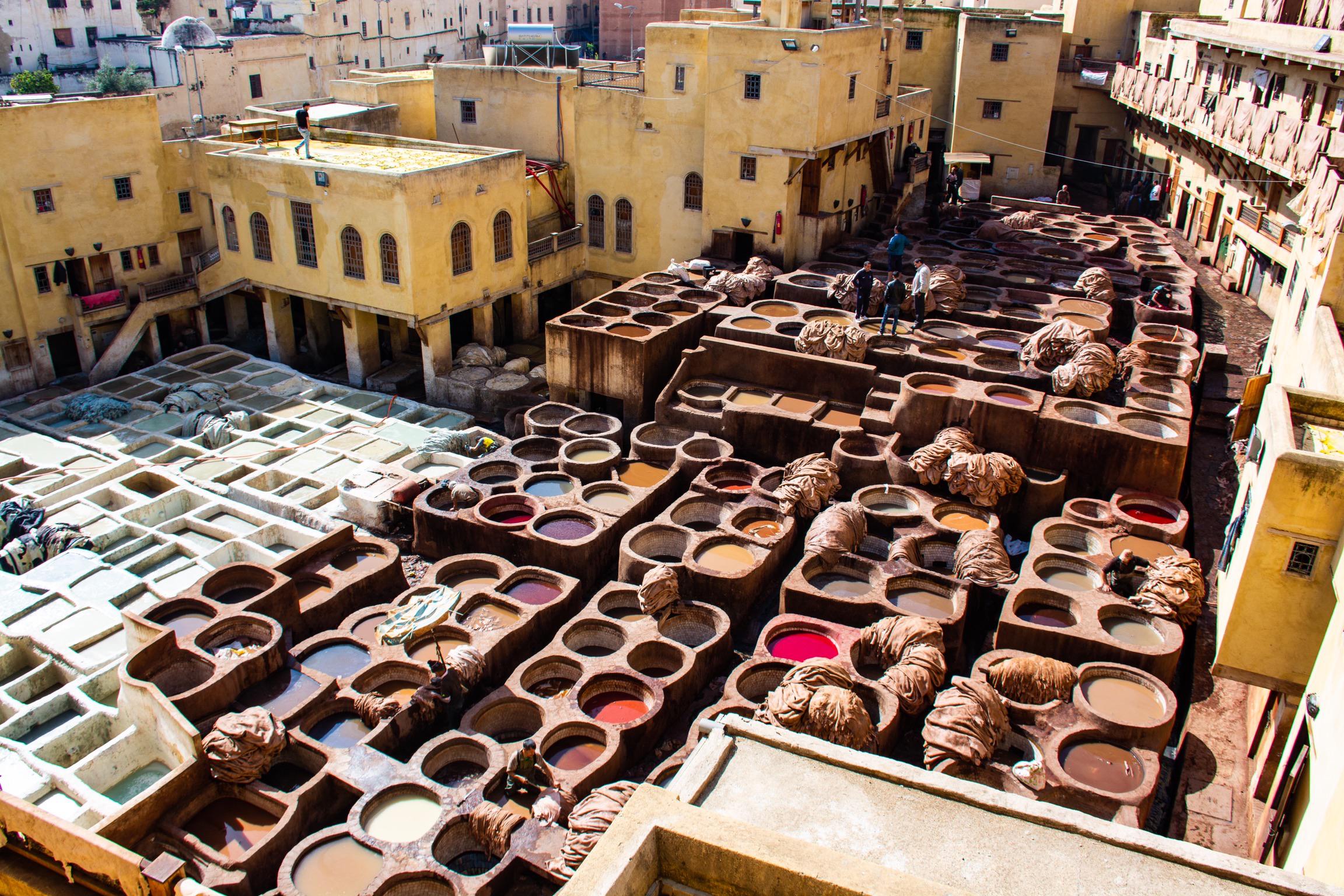 A vibrant tannery with large, round vats of colorful dyes and workers processing leather, surrounded by yellow buildings in an open courtyard.