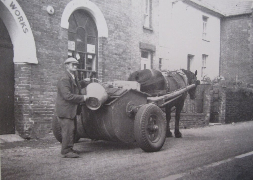 A man stands next to a horse-drawn cart on a street by brick buildings. The cart carries a large drum and other items. The scene appears old-fashioned and is in black and white.