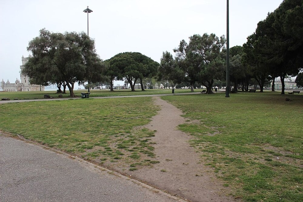 A dirt footpath cuts through a grassy park with scattered trees and lamp posts, leading toward distant historic buildings under an overcast sky.