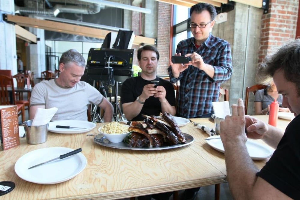 Four men sit around a table with a large platter of ribs and a bowl of macaroni. Two men take photos of the food, while the others look at their phones. The setting appears to be a casual restaurant.