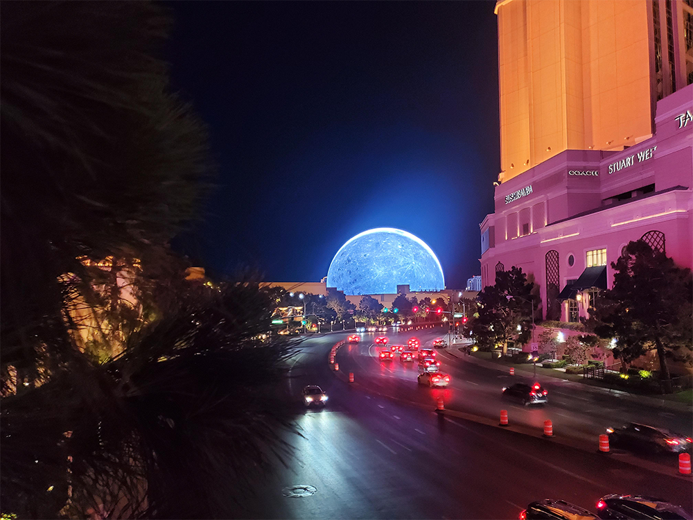 A night view of a busy Las Vegas street with cars, hotels, and the brightly lit MSG Sphere in the background displaying a glowing blue image.