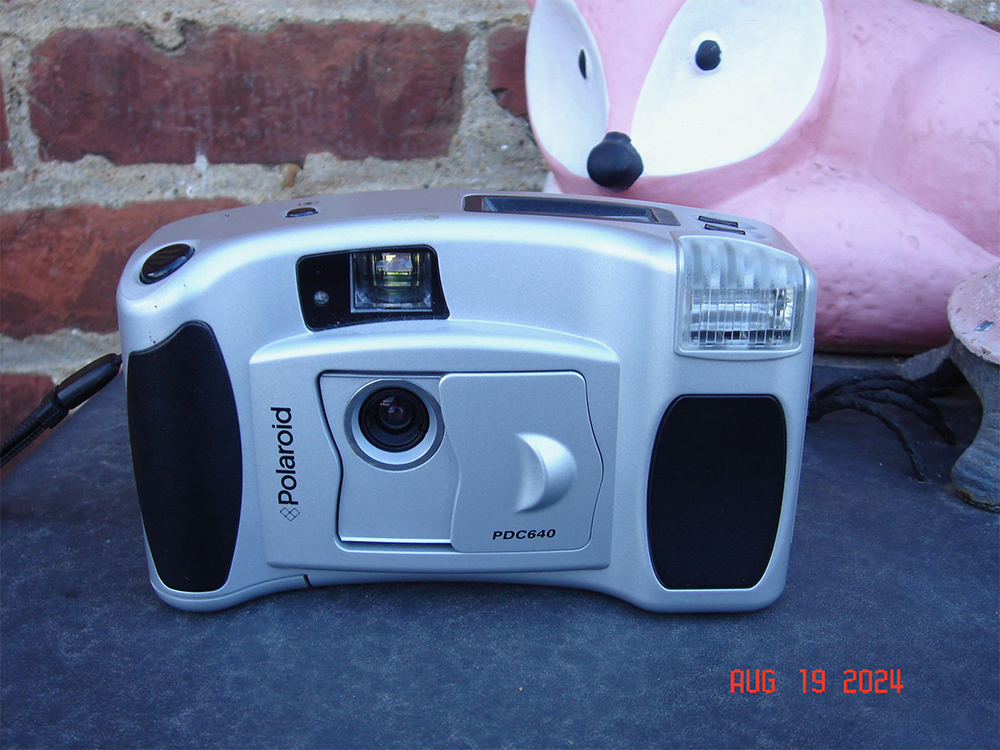A silver Polaroid PDC640 digital camera is on a blue surface, with a pink, fox-shaped object and a brick wall in the background. The date “AUG 19 2024” appears in red in the bottom right corner.