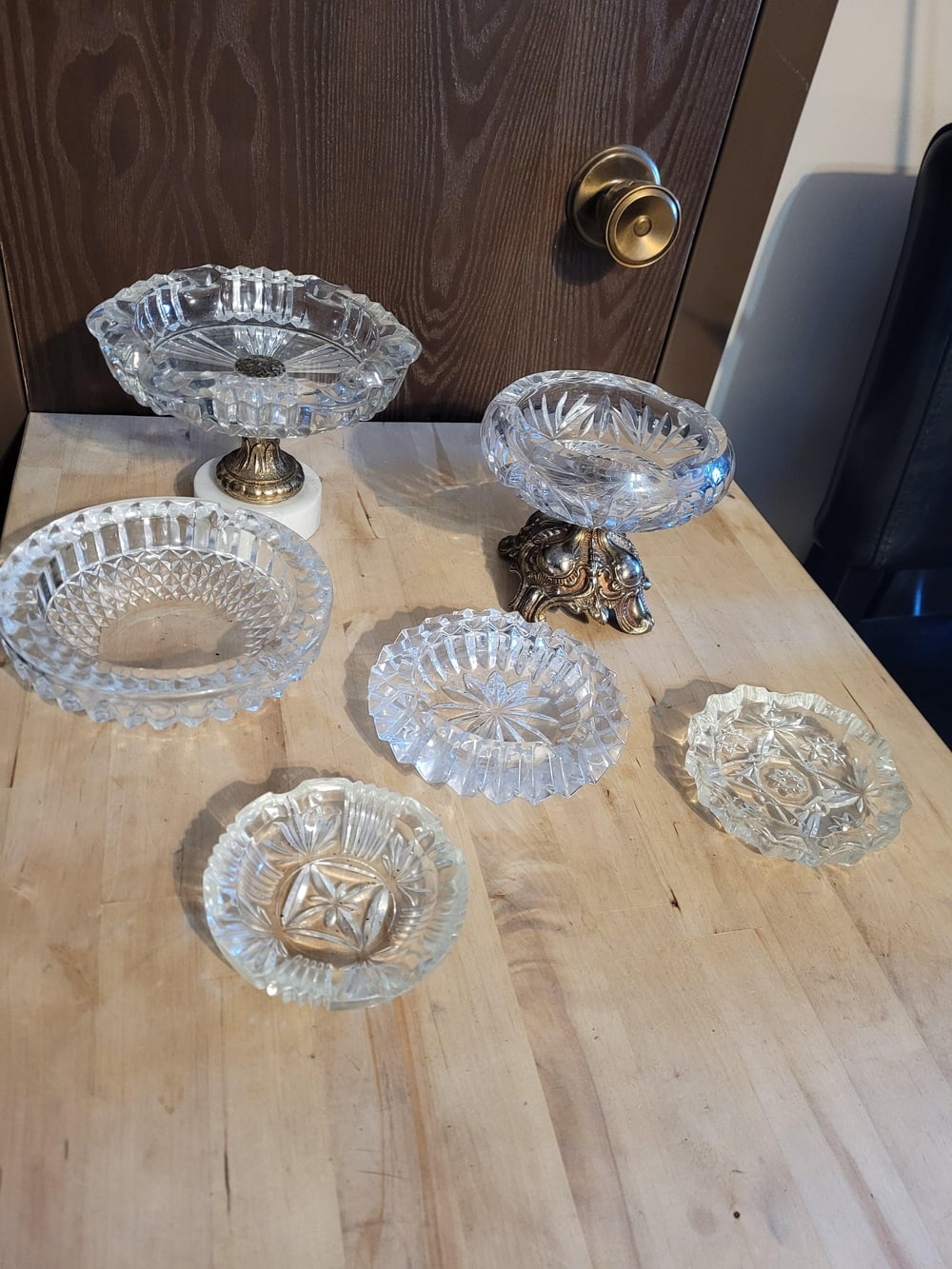 Seven vintage glass candy dishes and bowls with ornate designs are displayed on a wooden table in front of a brown door with a gold handle. Two dishes have decorative metal stands.