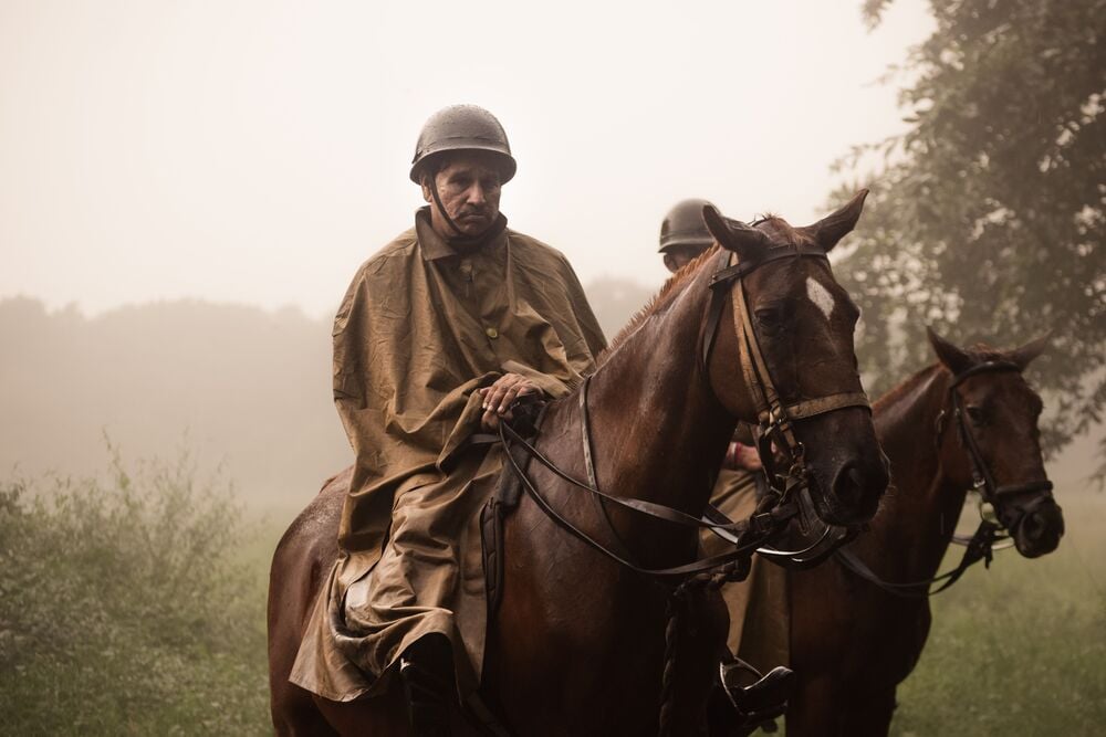 A person in a brown rain poncho and helmet rides a brown horse through a misty field, with another rider partially visible behind.