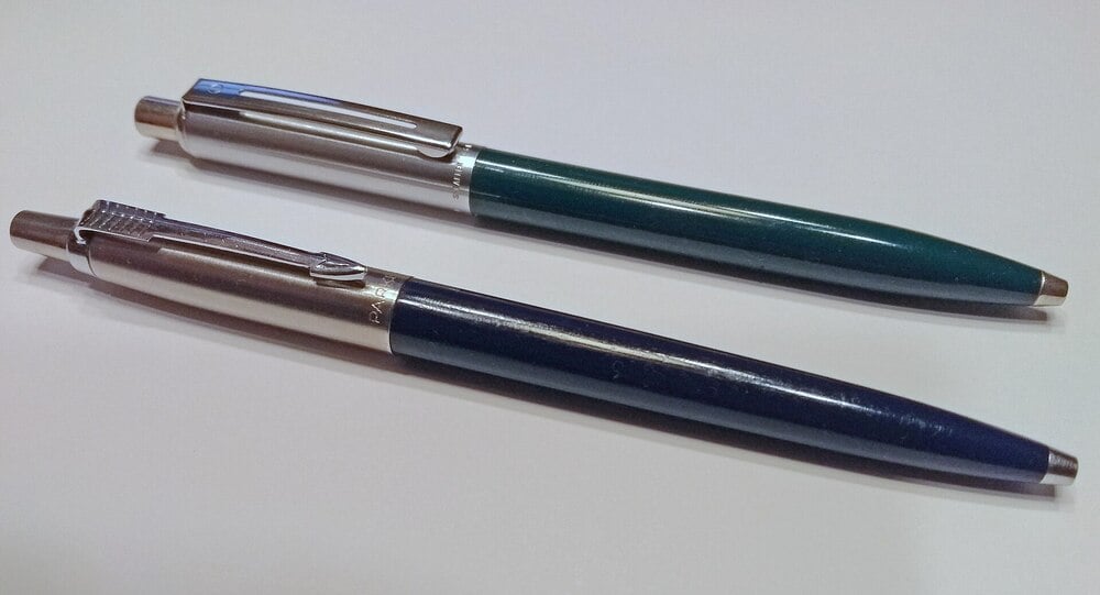 Two retractable ballpoint pens with metallic clips and tips lie parallel on a white surface. One pen has a dark green barrel, and the other has a dark blue barrel.
