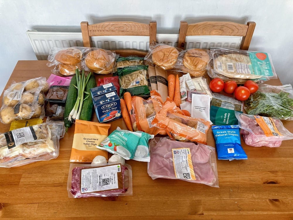 A wooden table with various groceries including bread rolls, vegetables, cheese, pasta, milk, meat, tomatoes, herbs, and packaged foods. Three wooden chairs are behind the table.