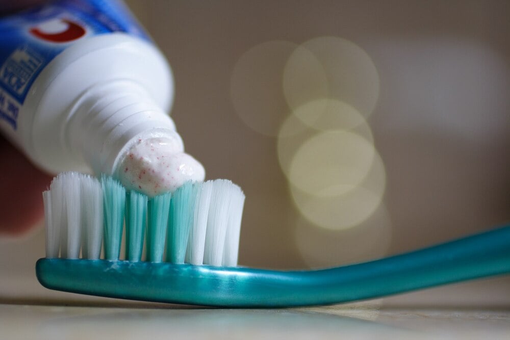 A close-up of toothpaste being squeezed onto the bristles of a turquoise toothbrush, with a blurred background.
