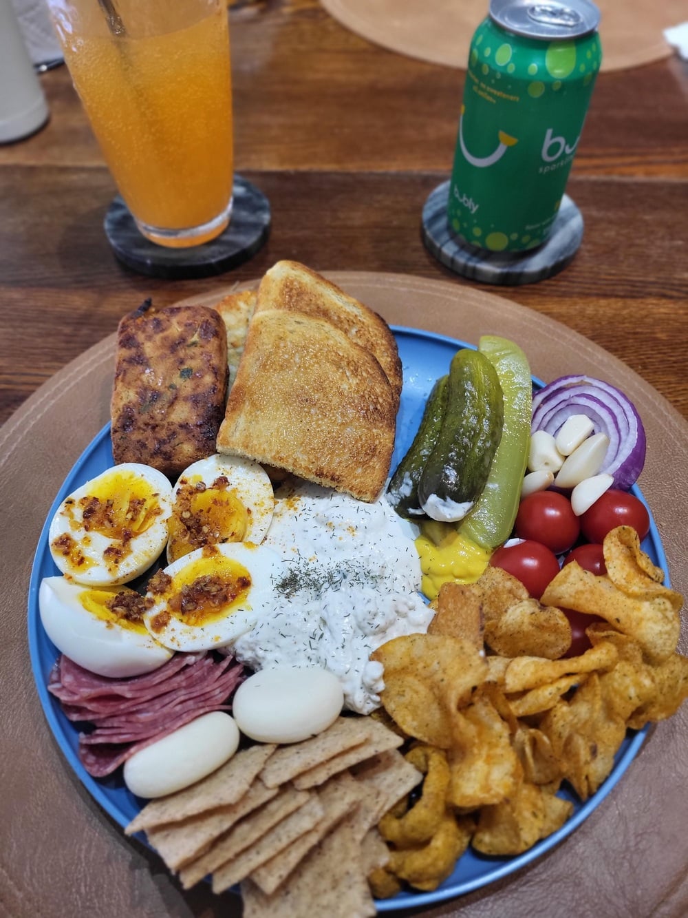 A blue plate with bread, seasoned boiled eggs, salami, cheese, chips, tomatoes, pickles, red onion, dip, and crackers. An orange drink and a green can are in the background on a wooden table.